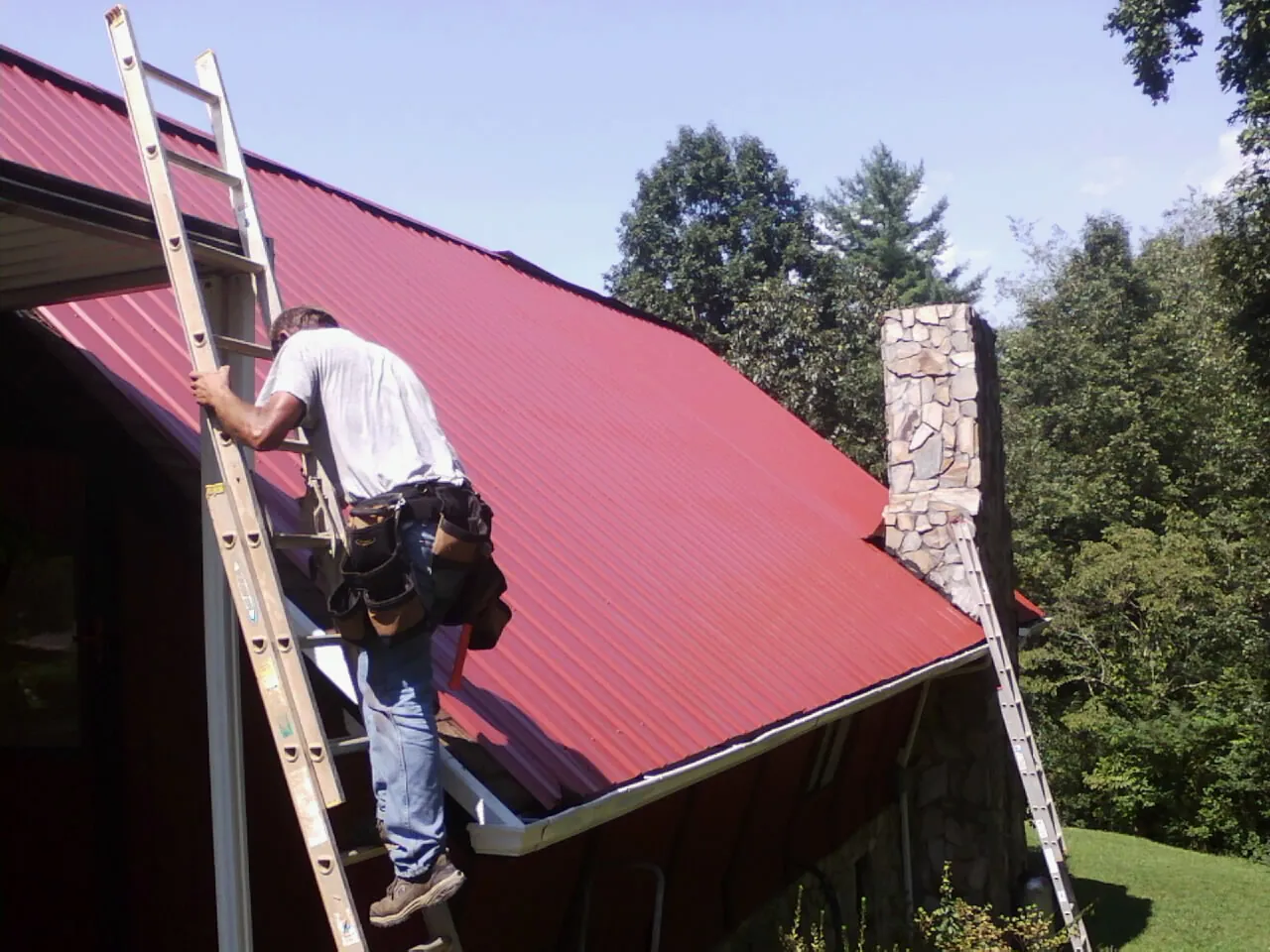 Roofer on a ladder beside a red metal roof and stone chimney — Soffit & Fascia Repair by Roofing Brothers in Grand Forks